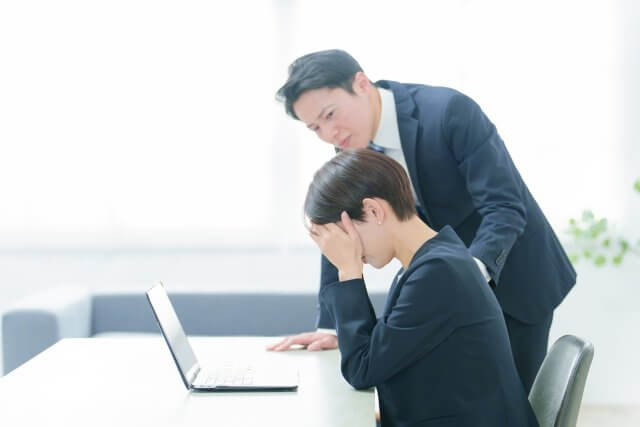 オフィスで、ノートパソコンの前で顔を覆いストレスを感じている様子のビジネスウーマンと、傍らに立つ男性の同僚（または上司）の画像。