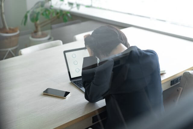 オフィスで、ノートパソコンの画面にグラフが表示されているのを見ながら、頭を抱えてストレスを感じている女性ビジネスパーソン。手前にはスマートフォンが置かれている。