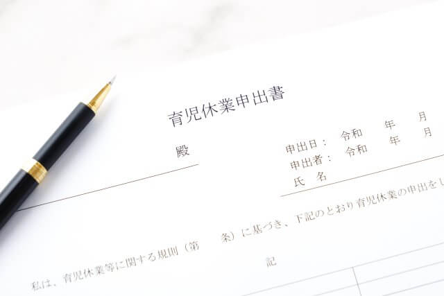 白い背景に「育児休業申出書」と書かれた書類が置かれ、その横に黒いペンが添えられている画像。育児休暇の申請手続き、法的な権利、ワークライフバランス、雇用主と従業員の関係性といった概念を表現しています。