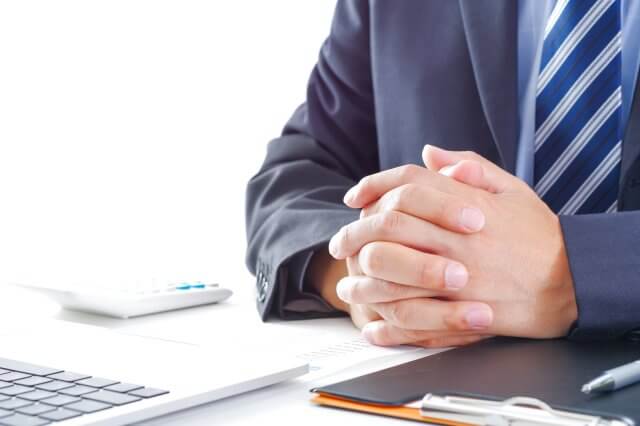 デスクで手を組んで座るビジネスマンのクローズアップ。ノートパソコンと書類が置かれたビジネスシーン。会議、商談、集中、真剣な態度を表現。